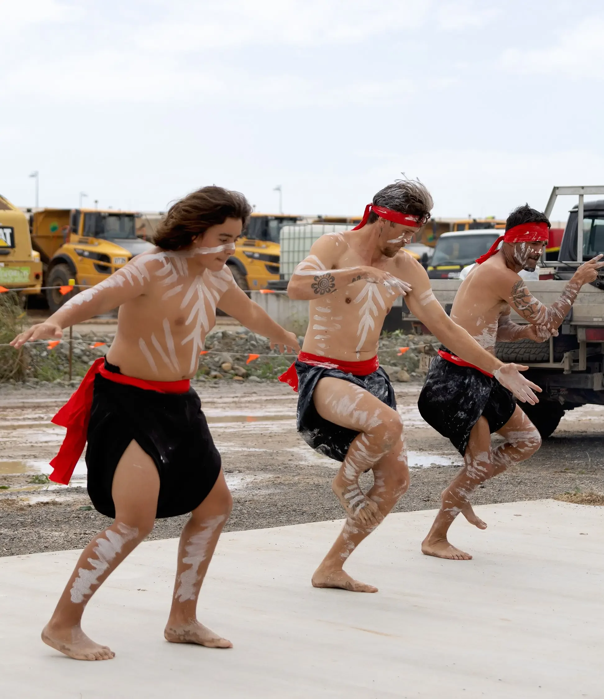 Four people participating in a traditional first nations dance, with painted bodies and red sashes, outdoors near construction vehicles.
