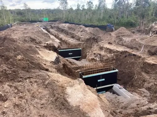 Construction site with large black concrete structures installed in trenches, surrounded by dirt and soil, with trees in the background.