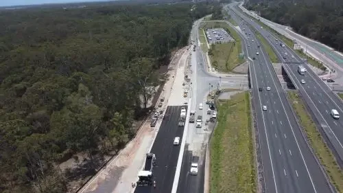 Aerial view of a highway with multiple lanes and vehicles, adjacent to a wooded area, with construction work on one side.
