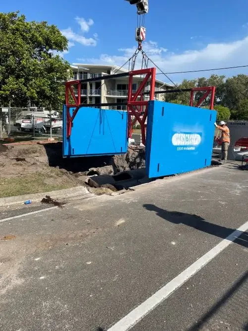 Construction site with a large blue and red metal barrier being lowered into a large hole in the ground, with a worker in a hat standing nearby.