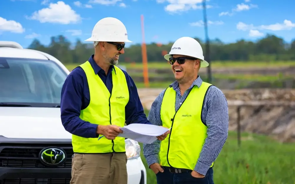 Two men wearing hard hats and yellow hi-vis vests review plans, smiling at each other, with a white vehicle and construction works in the background