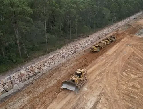 Construction site with bulldozers leveling the ground, surrounded by trees and a stone retaining wall.