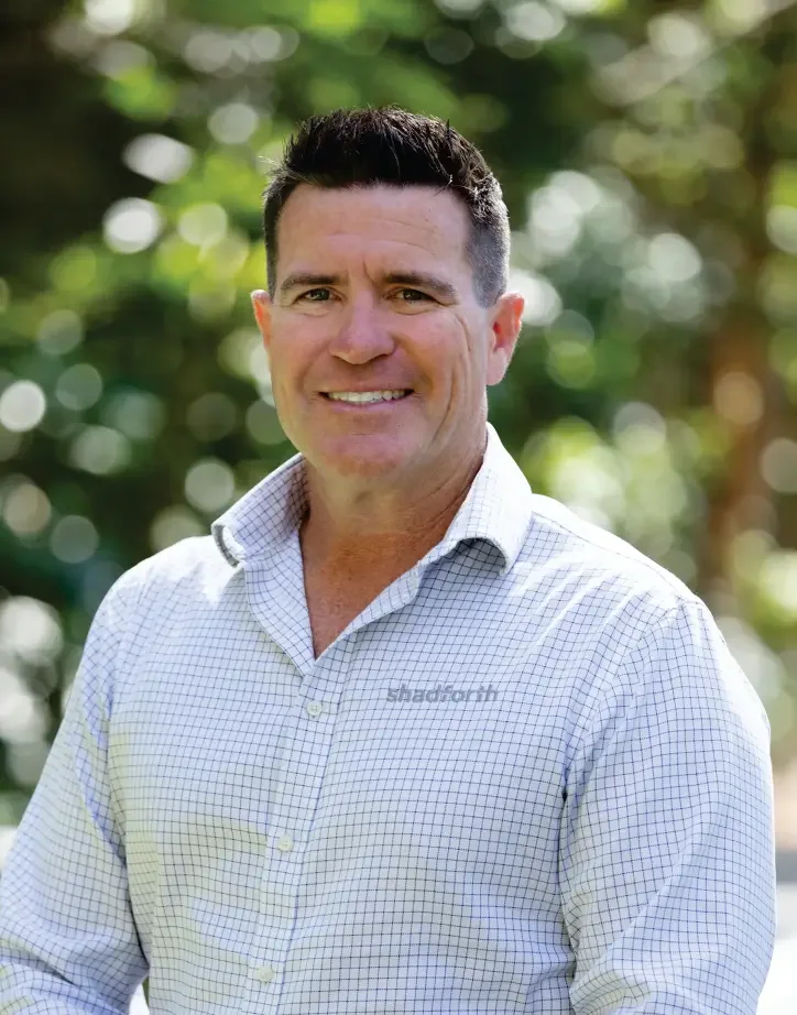 Company Director Ray Shadforth smiling outdoors, wearing a light checked shirt with the word 'Shadforth' embroidered on the shirt.