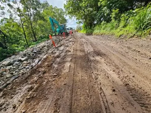 A dirt construction road with an excavator and orange cones on the side, surrounded by green trees.
