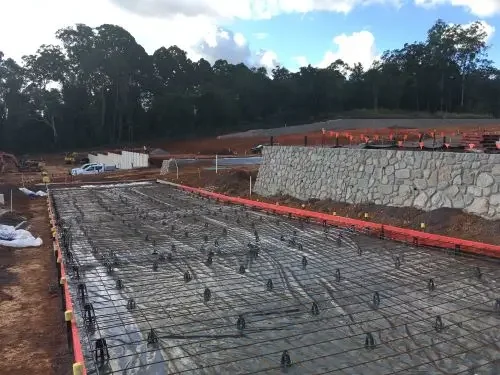 Construction site with rebar framework for a concrete slab, orange safety barriers, and a stone retaining wall, with trees and sky in the background.