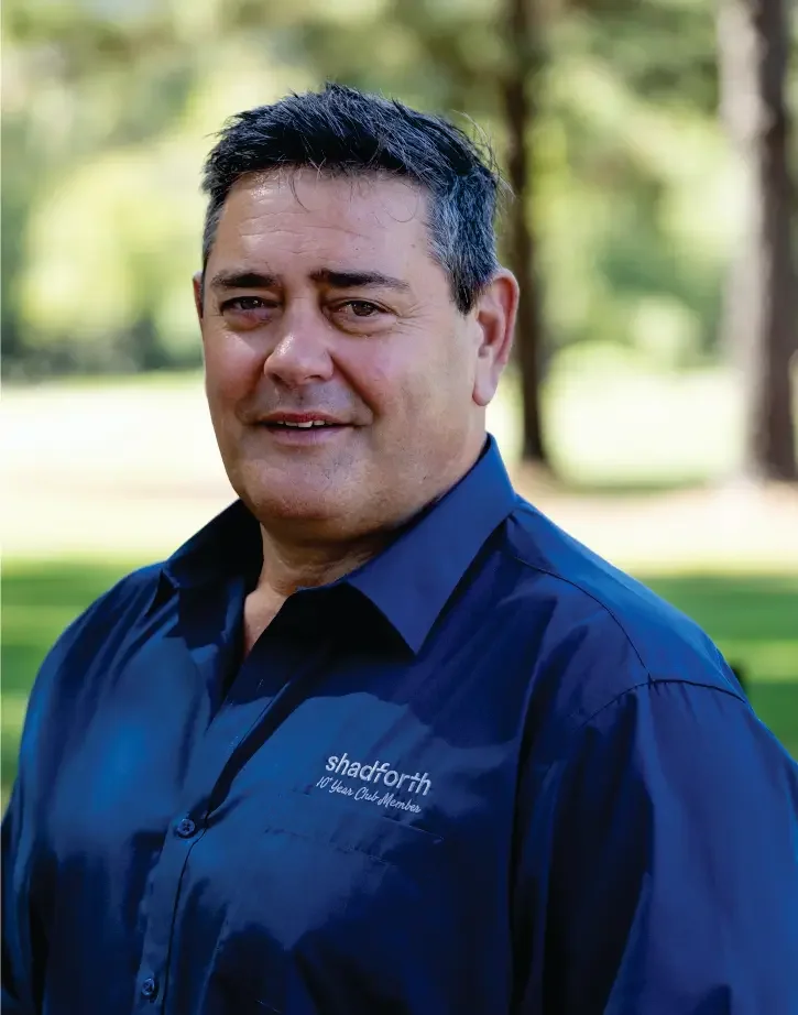 Chief Risk Officer Glen Panoho outdoors wearing a navy blue shirt with the Shadforth logo on the chest, with a blurred background of trees and grass.
