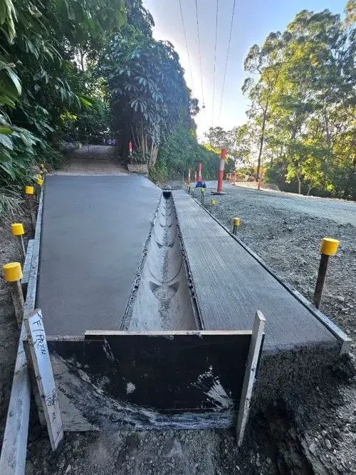 Newly constructed concrete pathway with a drainage channel in the middle, bordered by black and yellow safety barriers, on a road with trees and clear sky.