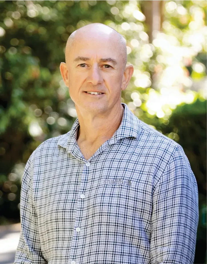 Executive Manager, Brad Moses in a checked shirt standing outdoors with greenery and sunlight in the background.