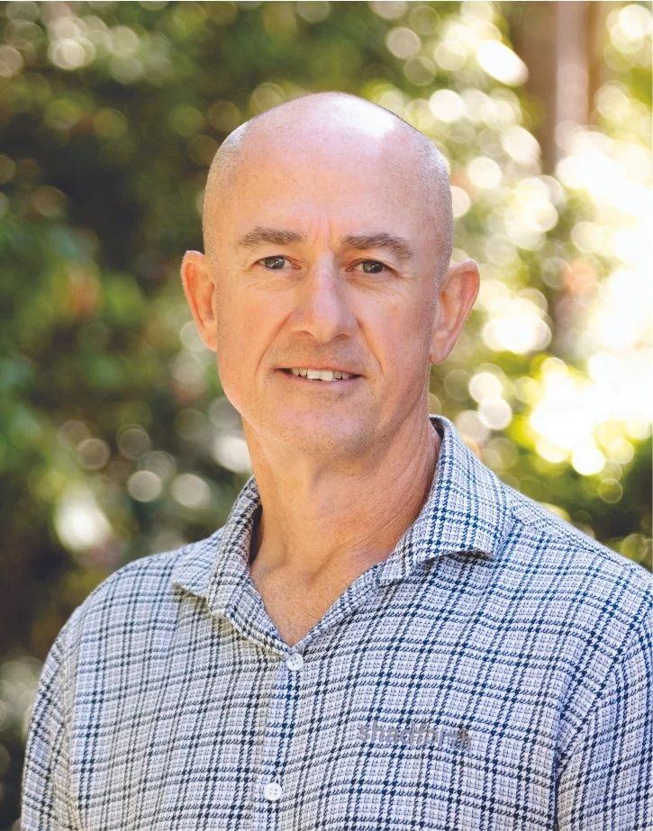 Executive Manager, Brad Moses in a checked shirt standing outdoors with greenery and sunlight in the background.