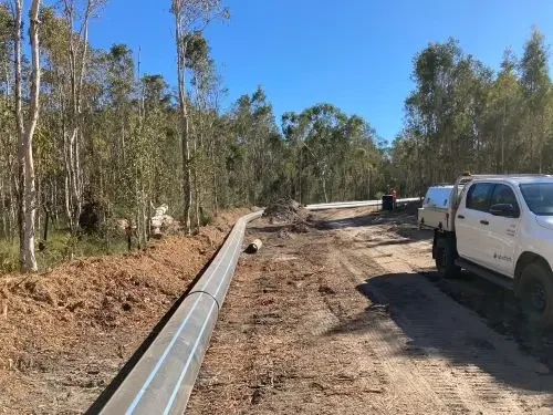 A construction site with a pipe laid along a dirt road, surrounded by trees under a clear blue sky. A white Shadforth ute is parked on the side of the road.