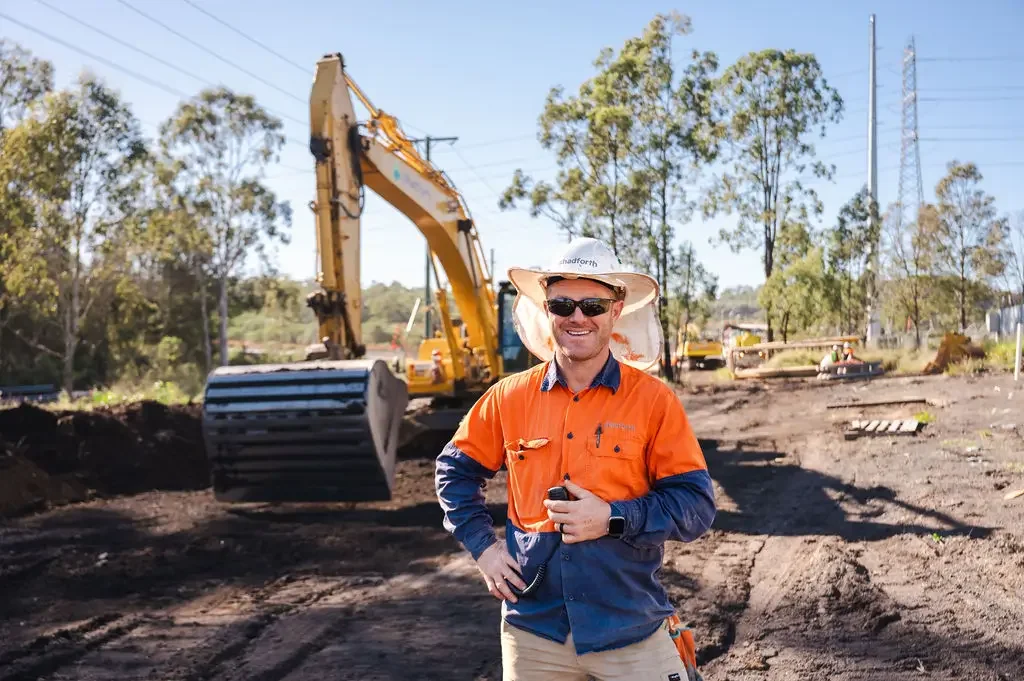 Construction worker standing in front of an excavator, smiling and holding a walkie-talkie, wearing a hard hat, sunglasses and an orange hi vis shirt.