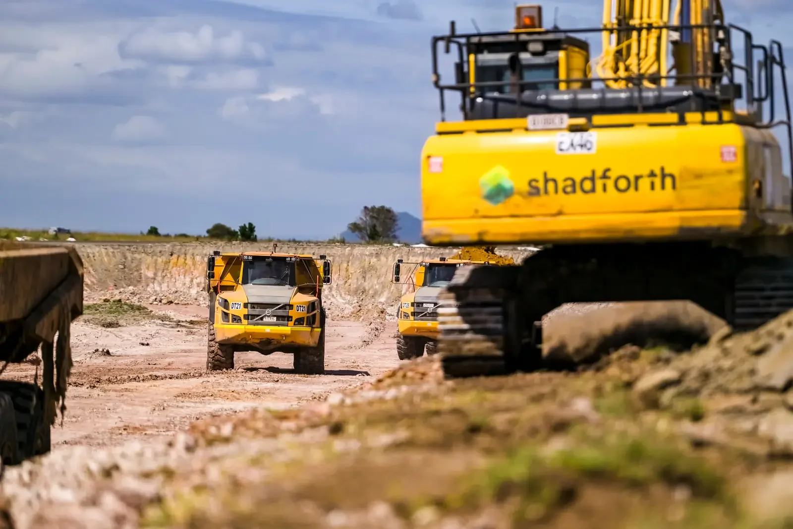 Construction site with large yellow excavator and two smaller yellow dump trucks driving on dirt.