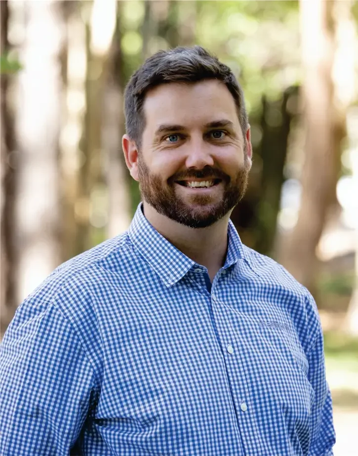 Executive Manager, Daniel Coonan wearing a blue and white checkered shirt, standing outdoors in a forested area with blurred trees in the background.