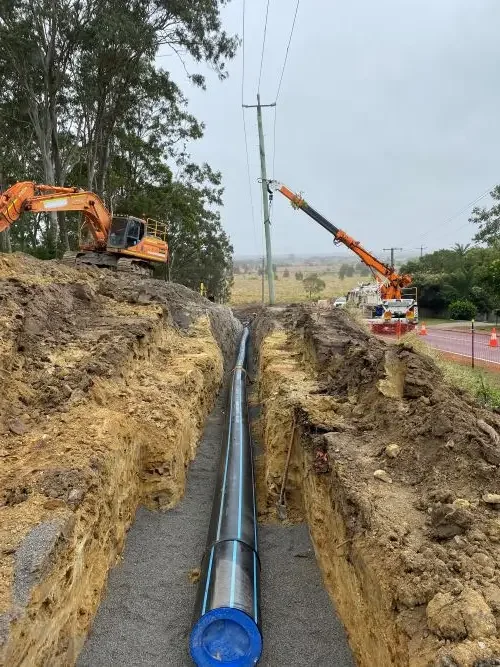 A construction site showing a large hole with a long black pipe installed inside. There is an excavator and a crane, and power lines above.