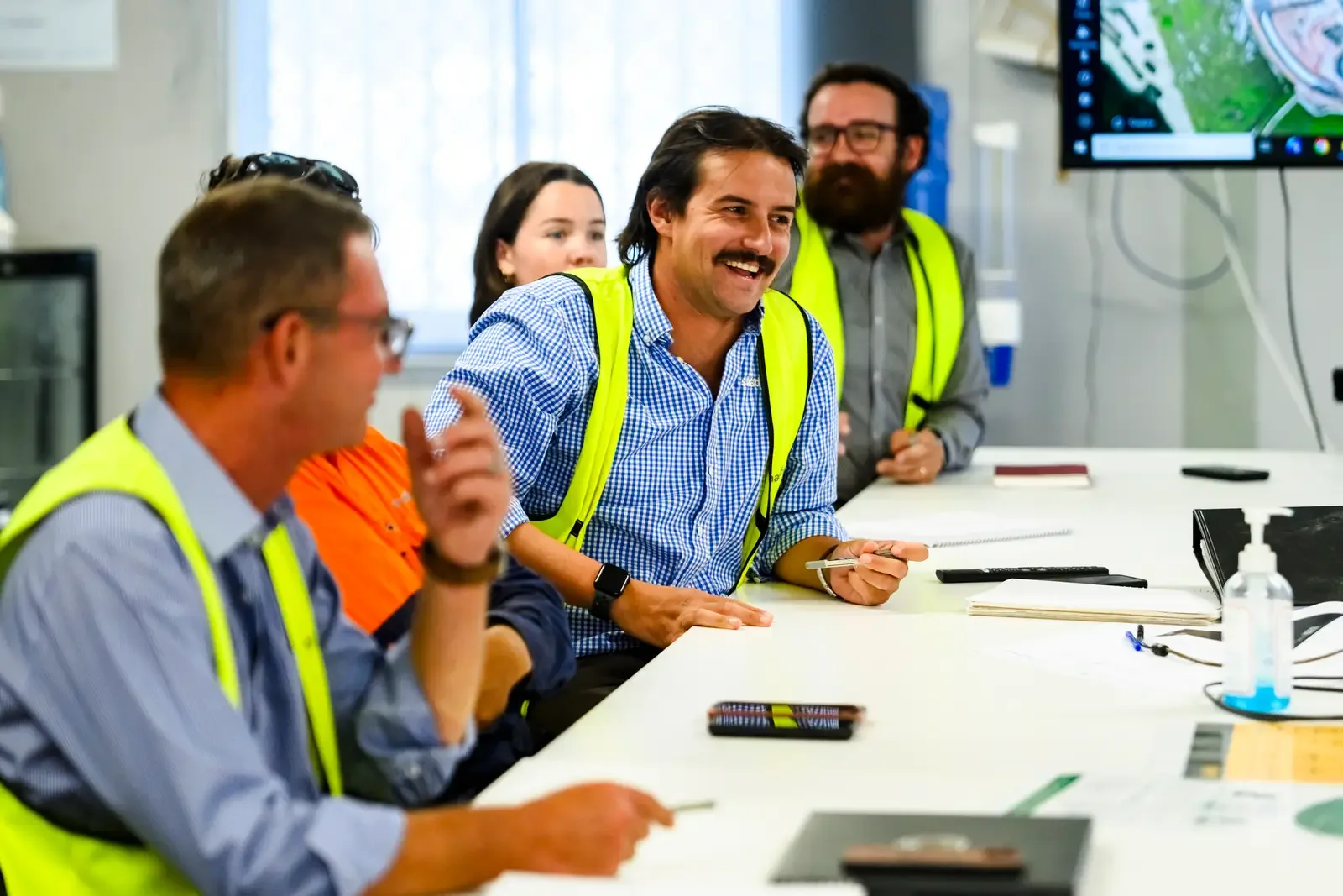 Five people sitting at a table in a site meeting talking. They are wearing hi vis vests.