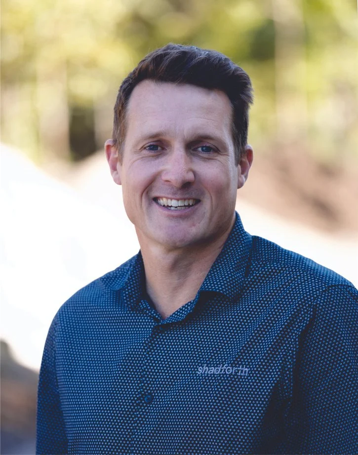 Executive General, Manager Michael Bishop wearing a blue patterned shirt outdoors with blurred green foliage in the background.