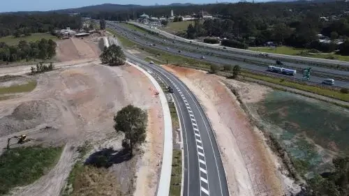 A newly constructed road and path with Bruce Highway and vehicles in the background, some trees and dirt on the sides.