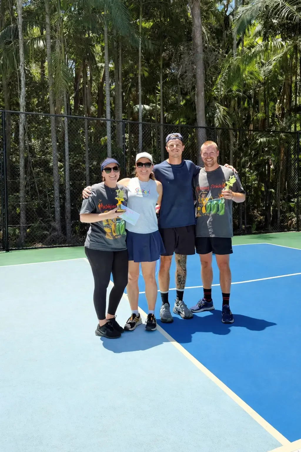 Four people standing on a tennis court holding trophies, smiling, with trees in the background.