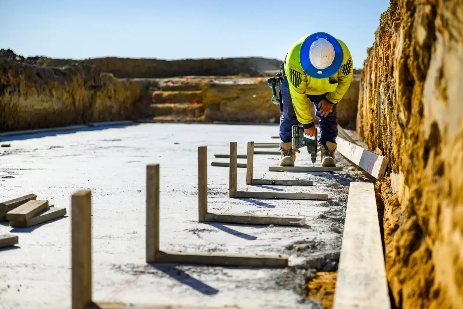 Construction worker measuring and preparing the foundation for a building, with wooden stakes and a level on a concrete surface.