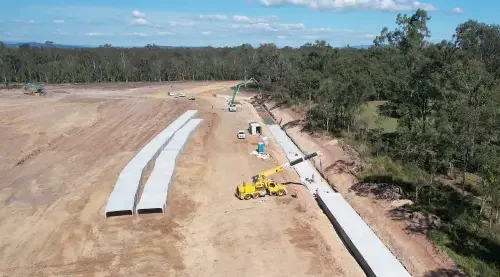 Construction site with large concrete beams, construction vehicles, and workers installing a barrier alongside a road in a rural area.