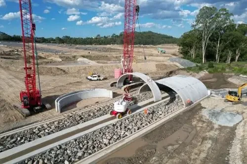 Construction site with large red cranes, a curved tunnel structure, and construction vehicles on a partially developed area with trees in the background.