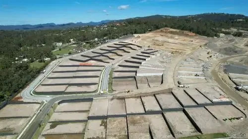 Aerial view of a suburban neighborhood under construction with cleared land, partially built homes, and surrounding greenery, with hills in the background.