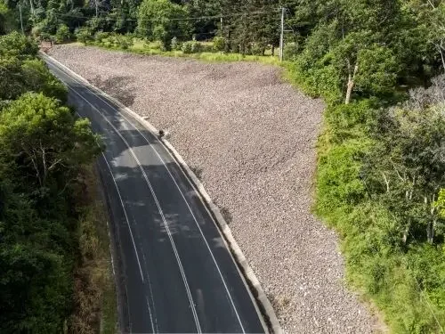 A winding paved road running through a wooded area with a gravel embankment on one side.