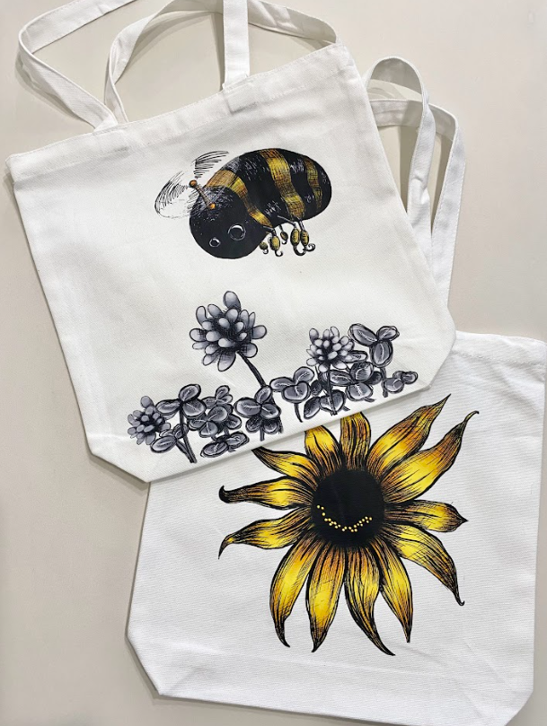 Reusable white grocery tote bags featuring multiple designs, including a smiling sunflower and flying bean artwork.