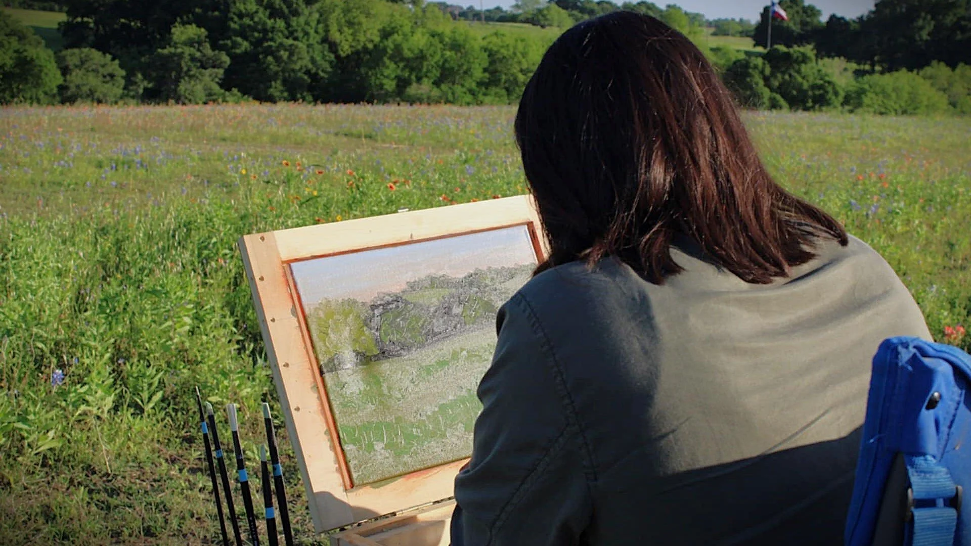 An artist painting plein air in a wildflower meadow, seated at a portable easel while overlooking rolling hills beneath an open sky.