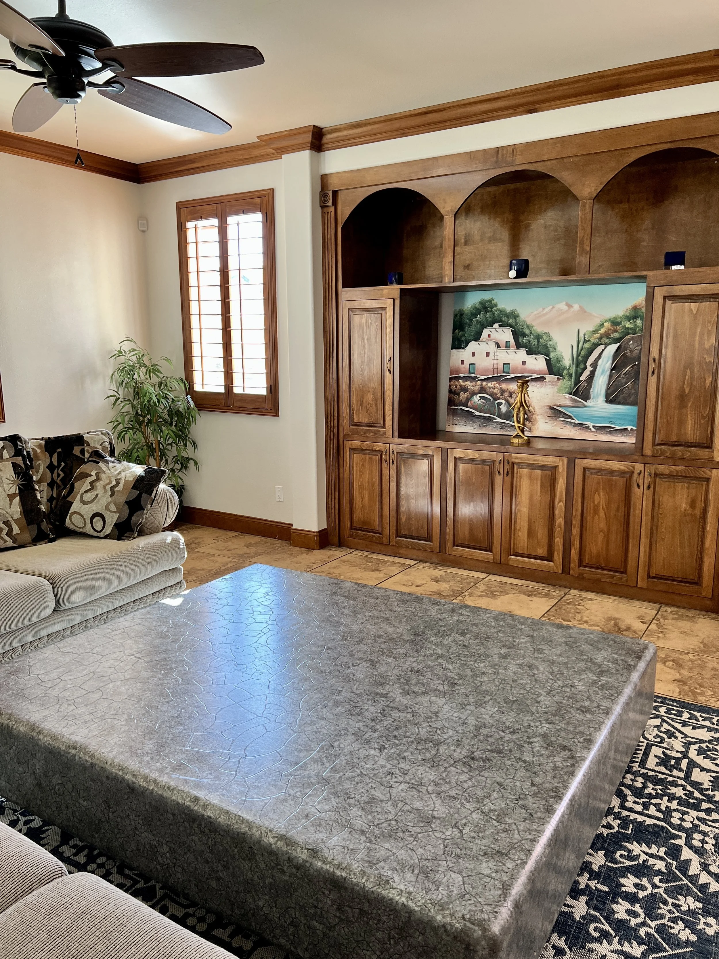 Living room with a beige sofa with patterned pillows, a large textured gray coffee table, a wooden entertainment center with a painted landscape scene, window with wooden shutters, and a ceiling fan.