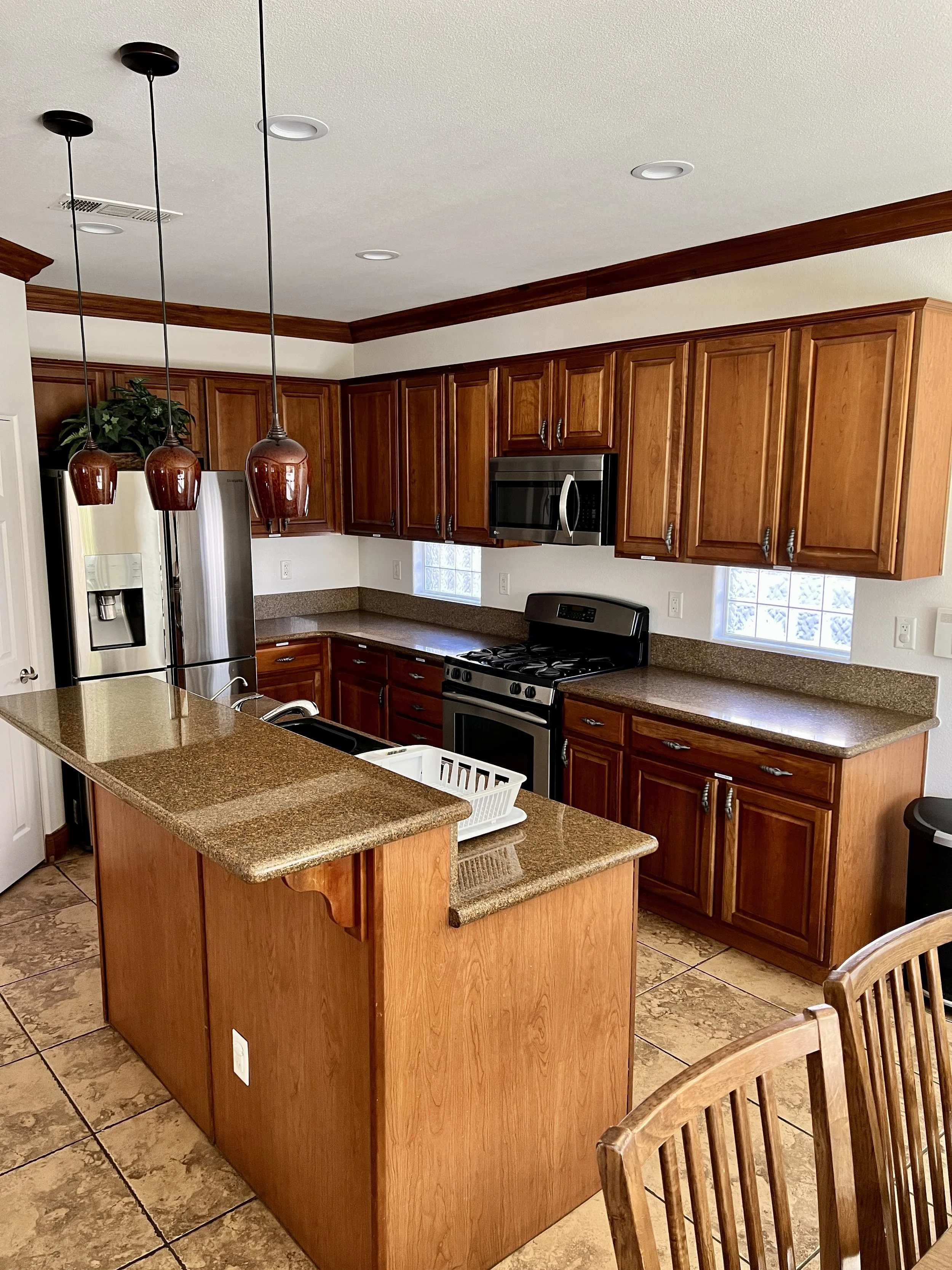 A kitchen with wooden cabinets, granite countertops, a stainless steel refrigerator, microwave, and stove. A kitchen island with a sink and hanging pendant lights. Tile flooring and small glass block windows.