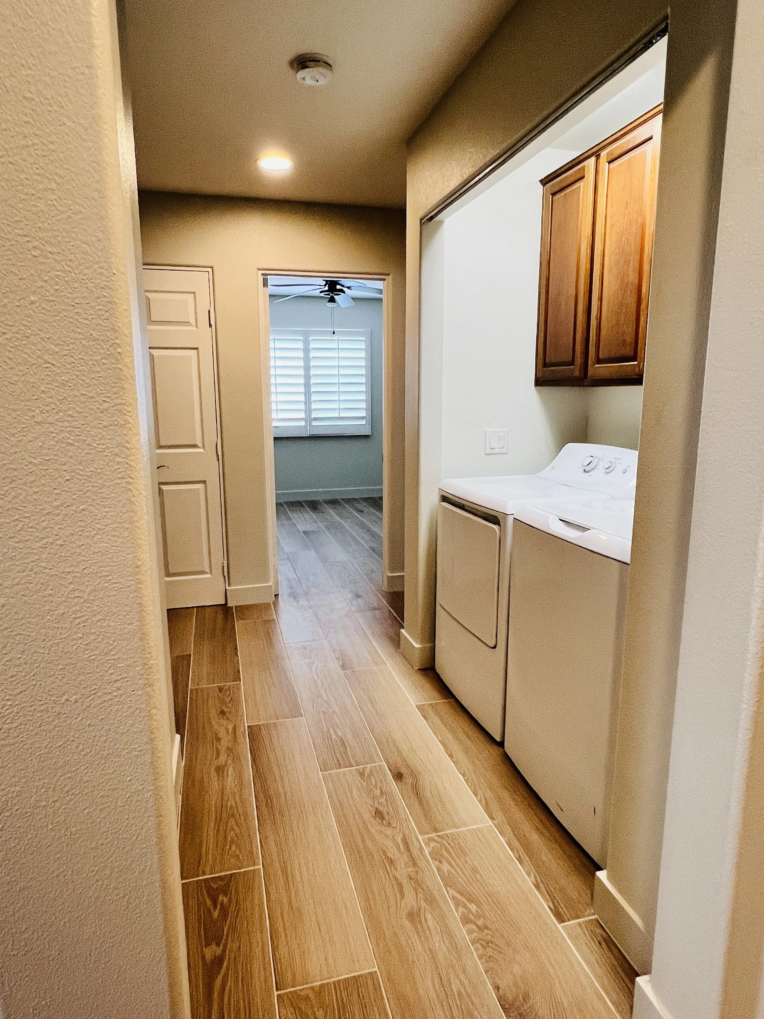 Laundry area with a white washing machine and dryer, wooden cabinets above, view towards a room with a window and blinds, hardwood floor.