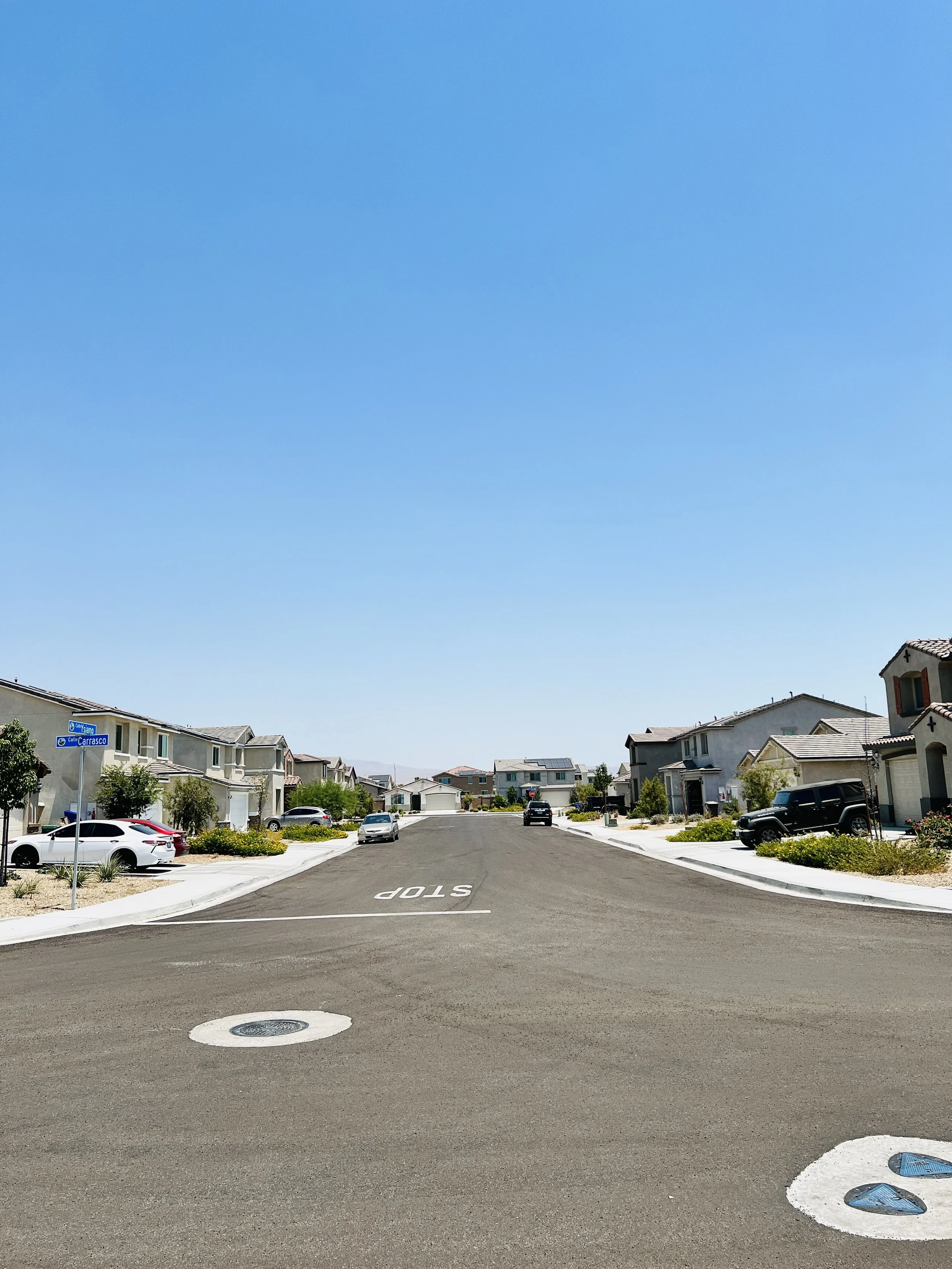 Residential street with houses, parked cars, clear blue sky, and stop sign at the intersection.