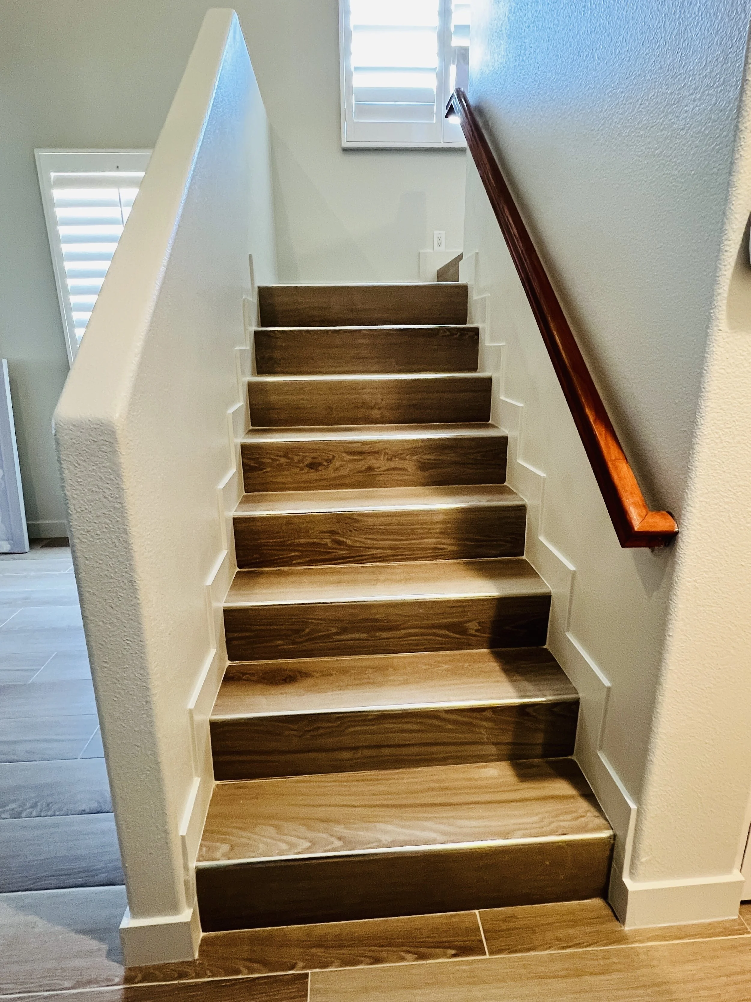 Wooden staircase with white risers, beige walls, and a wooden handrail on the right side. There are two windows with white blinds letting in natural light.