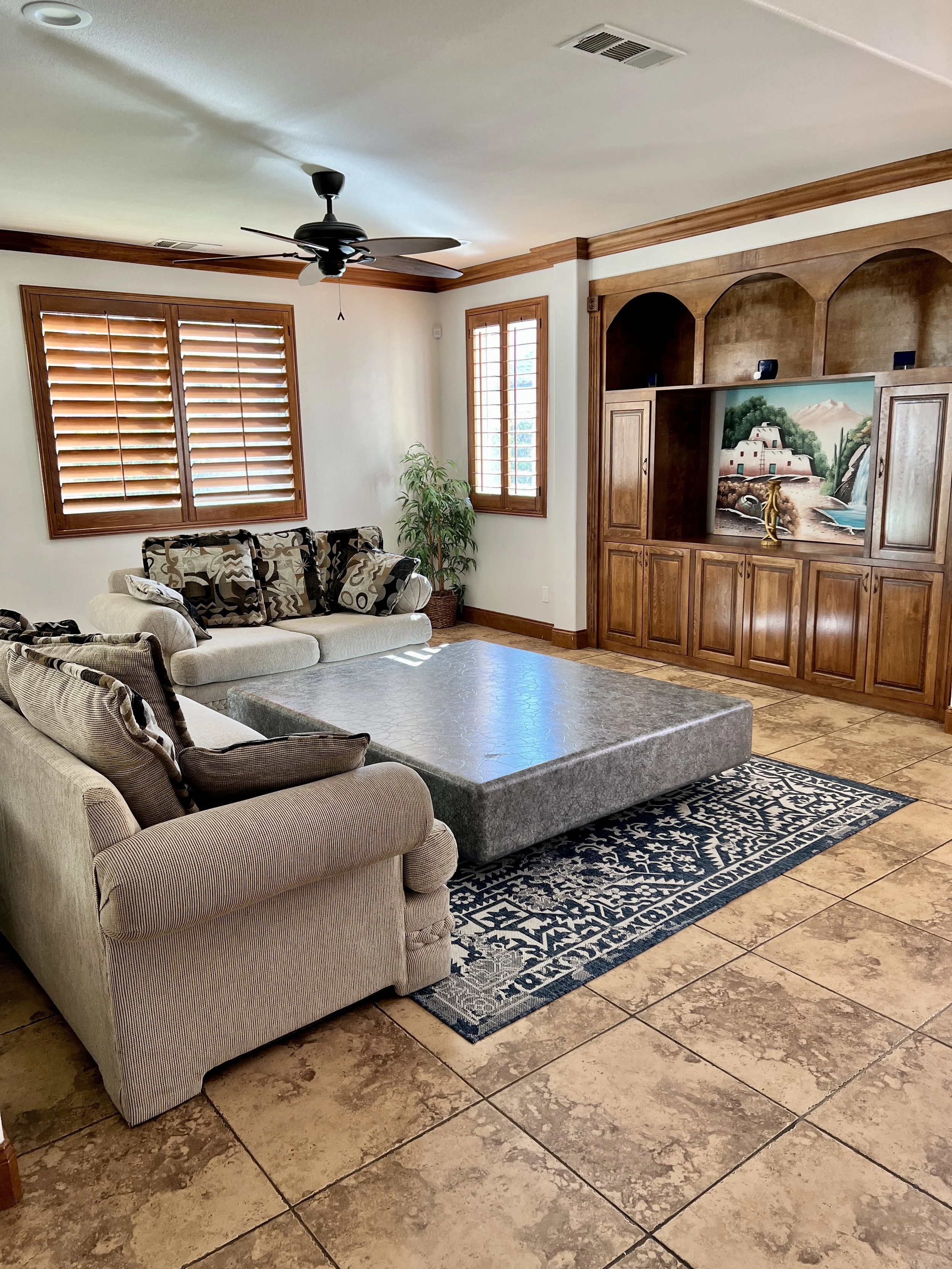 Living room with beige sofas, a large grey ottoman, a patterned rug, wooden window shutters, a ceiling fan, and a built-in wooden entertainment center with artwork and decorative items.