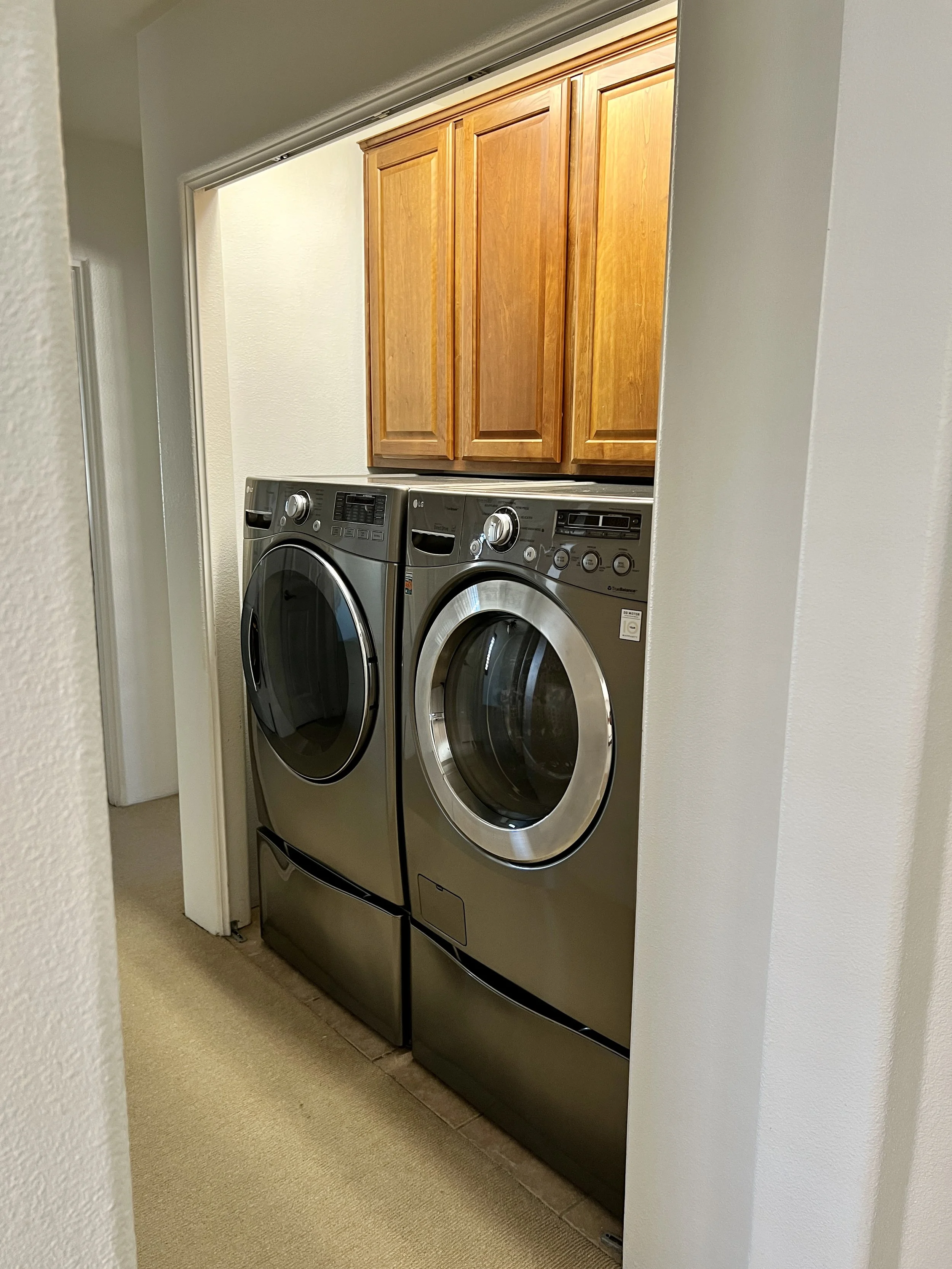 A laundry room with a front-loading washing machine and dryer, above which are wooden cabinets, in a beige-carpeted area.