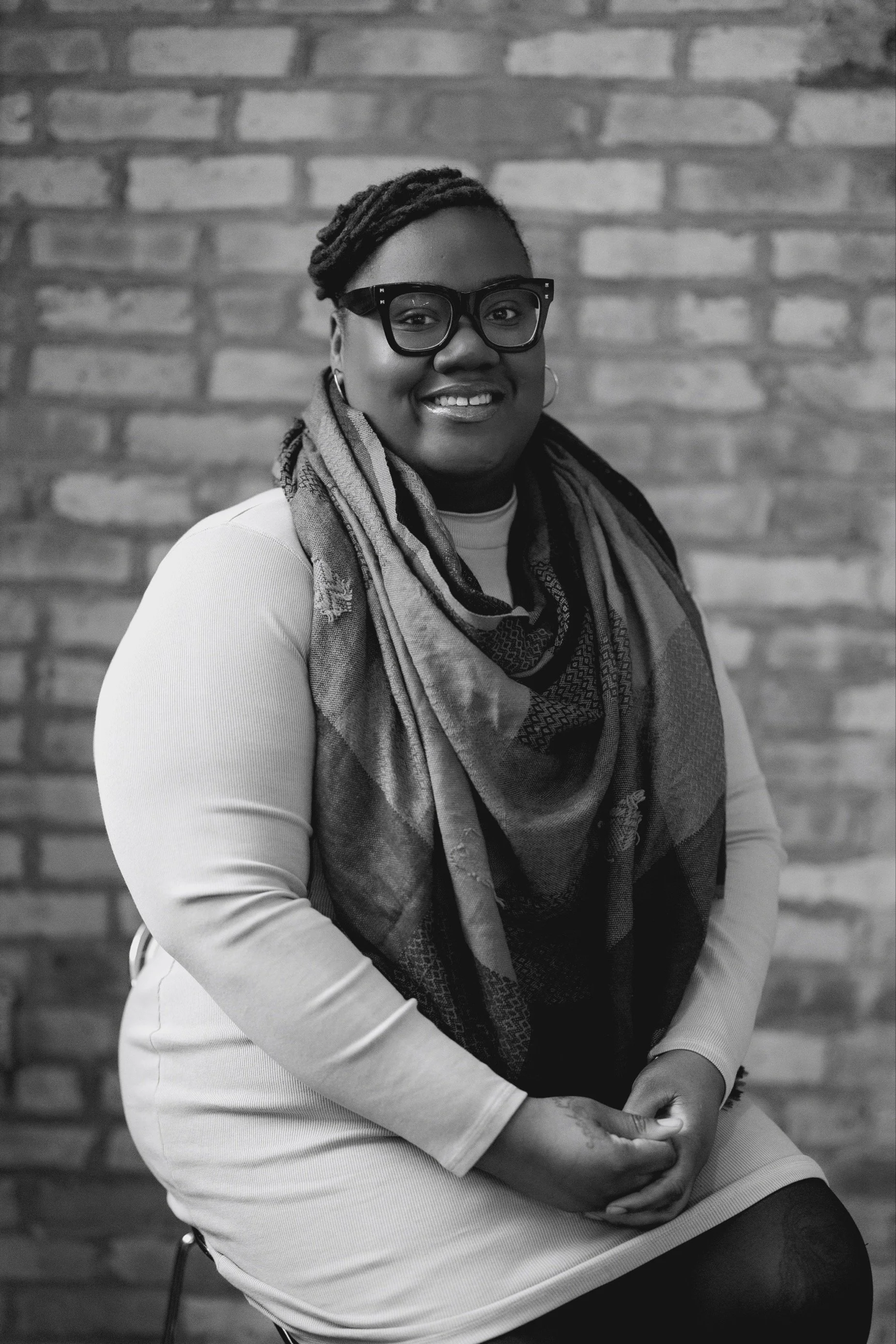 A woman smiling, wearing glasses, a light-colored dress, and a patterned scarf, sitting in front of a brick wall.