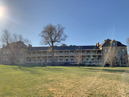 A large, multi-story building with a wrap-around porch, situated behind a grassy area with trees, under a clear blue sky.
