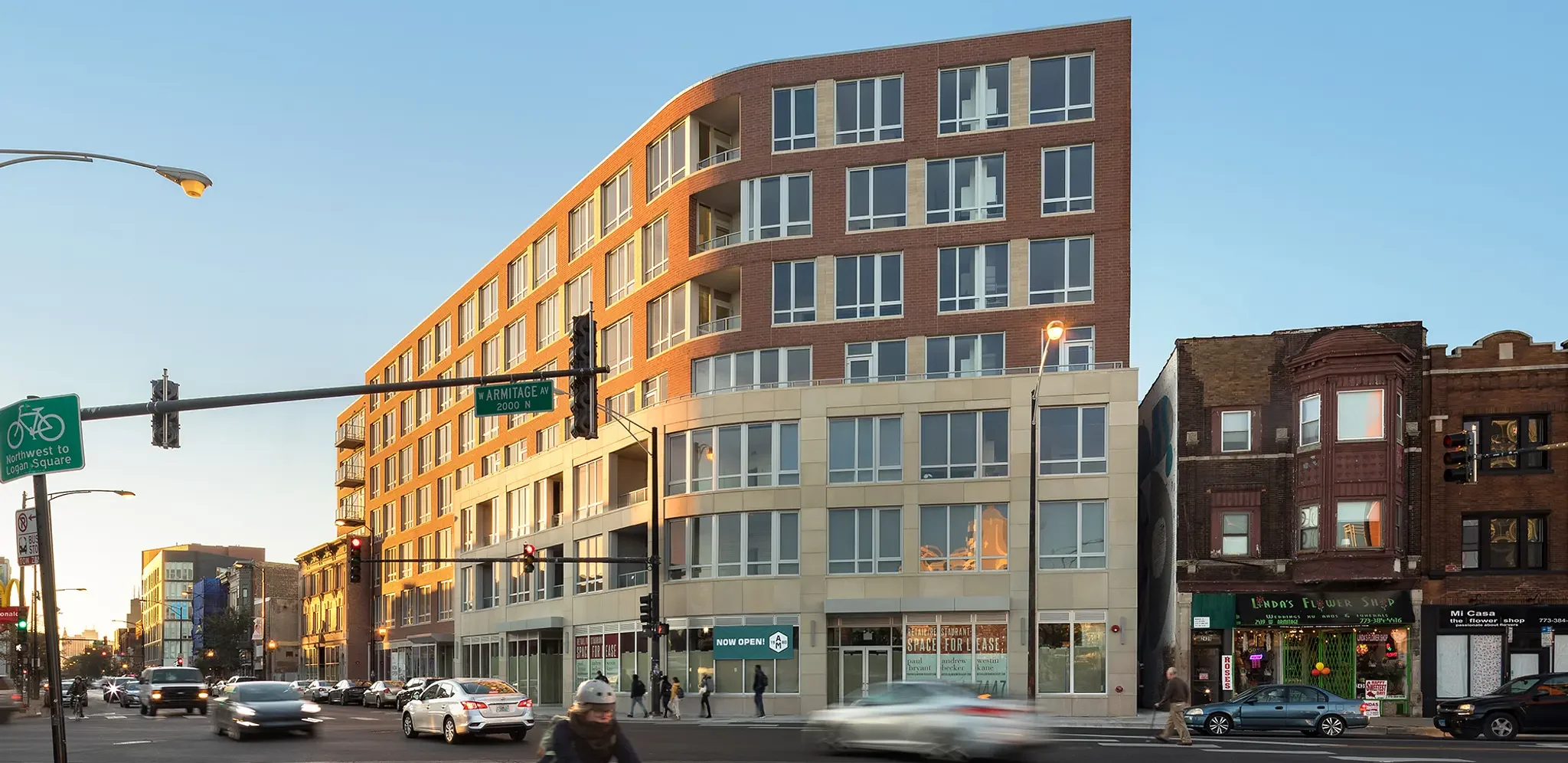 Modern multi-story building with large windows across the street from a busy intersection with cars and pedestrians