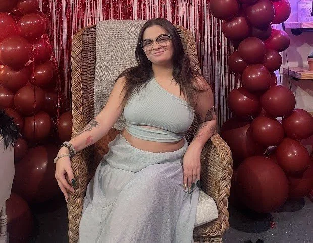 A woman with long dark hair and glasses sitting on a wicker chair, wearing a light gray crop top and matching long skirt, surrounded by red balloons and red curtain decorations.