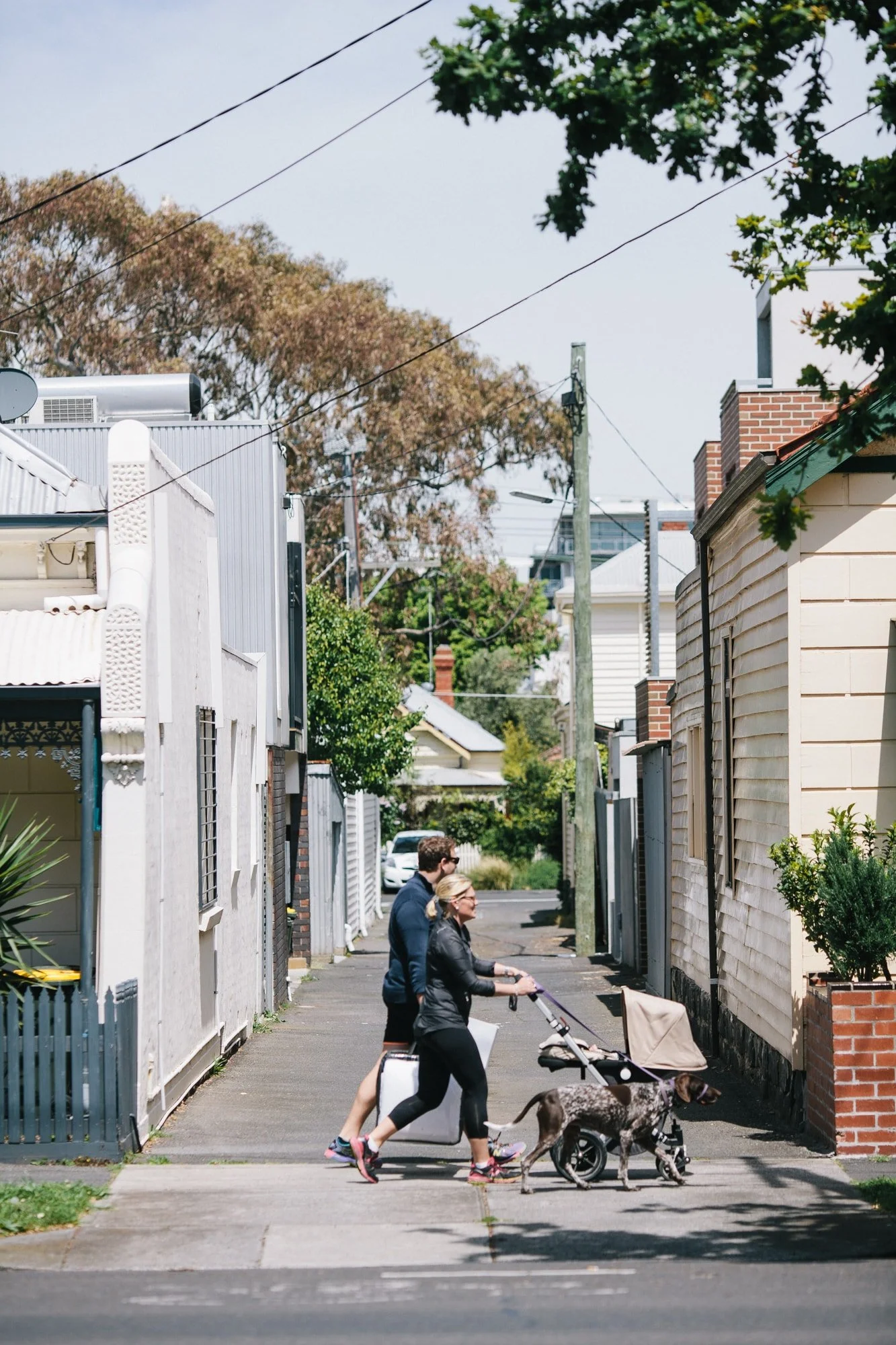 A man and woman walking down a footpath with a dog in a pram, pulling a suitcase, in the Richmond neighbourhood with houses and trees.
