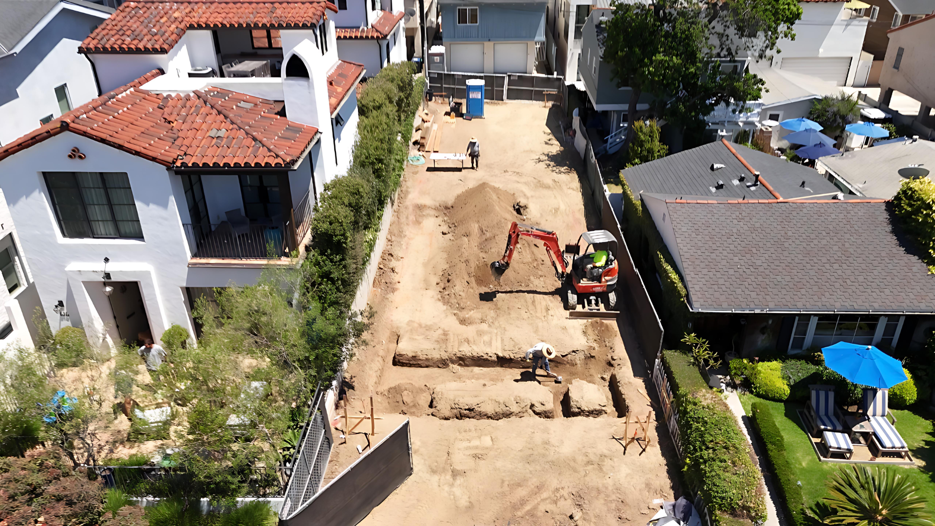 Aerial view of a residential construction site with workers, heavy machinery, and cleared land between houses with green lawns and outdoor furniture.