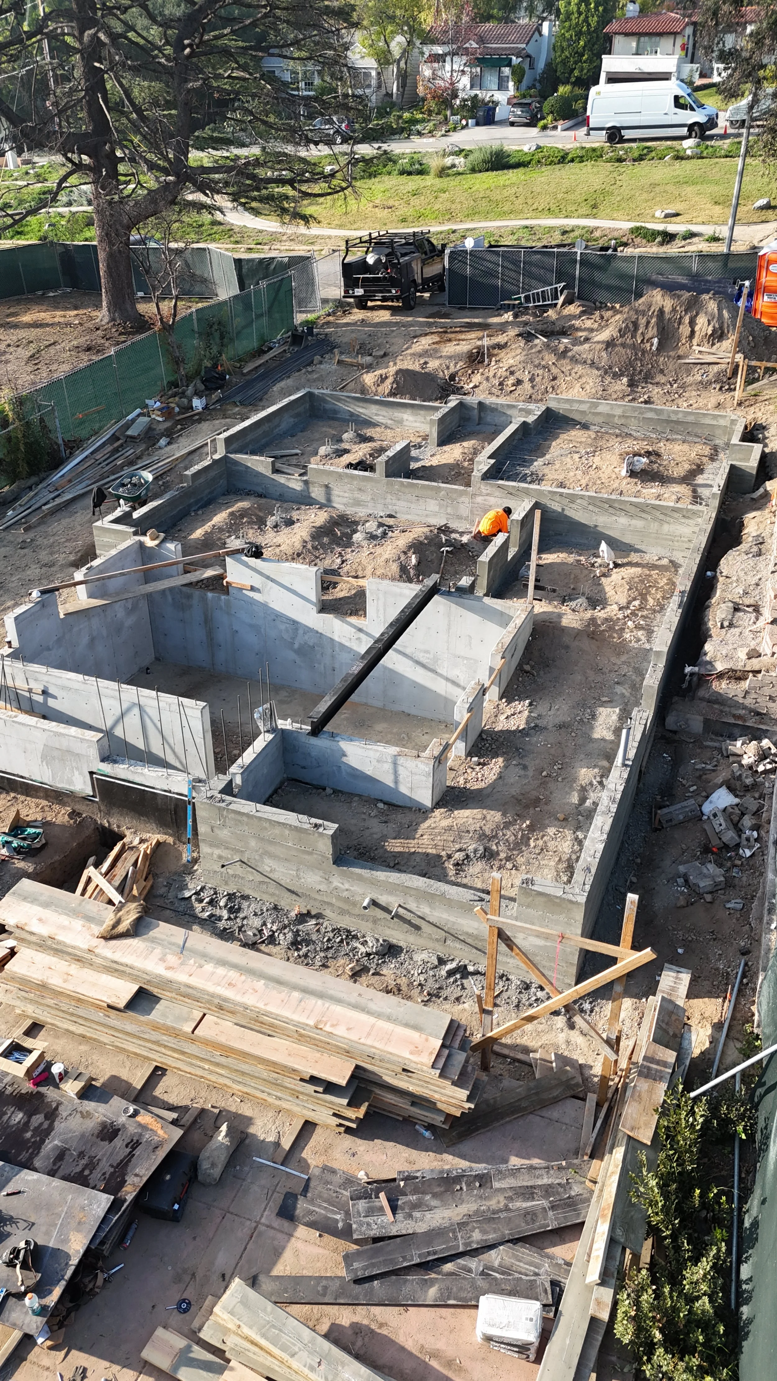 Construction site with concrete foundation and wooden framing, workers in orange safety vests, construction materials, and tools, with a residential neighborhood in the background.