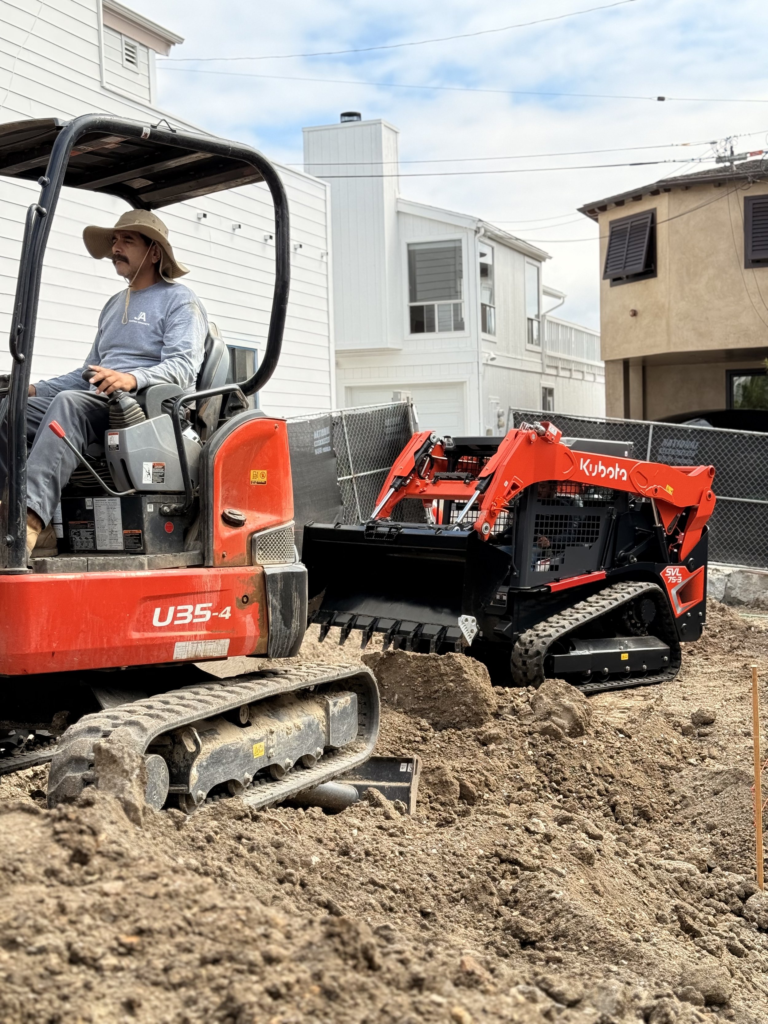 A man operating a compact excavator at a construction site in a residential area with houses in the background.