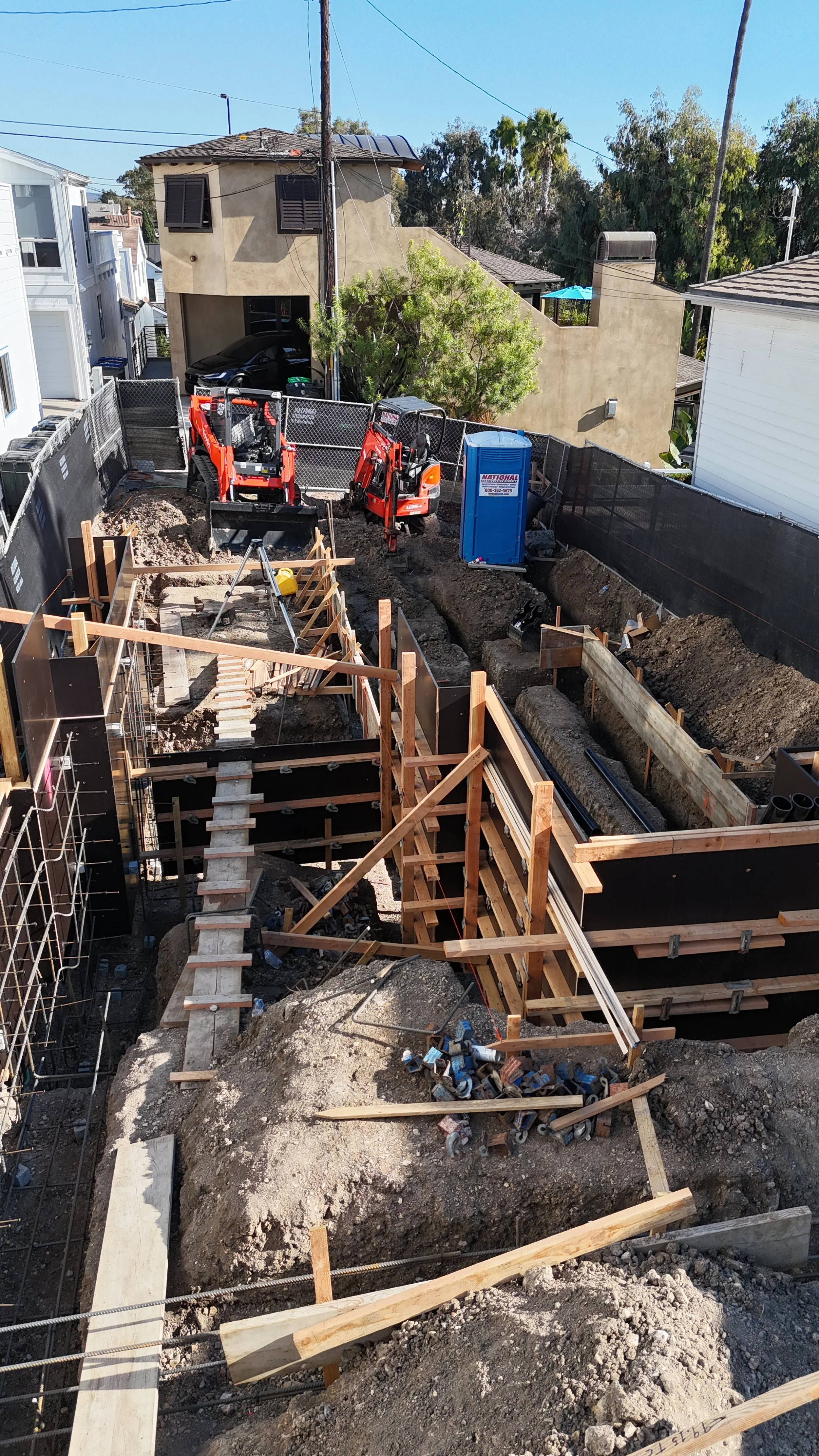 Construction site with wooden framework, digging trenches, heavy machinery, and construction materials for building foundation in a residential neighborhood.