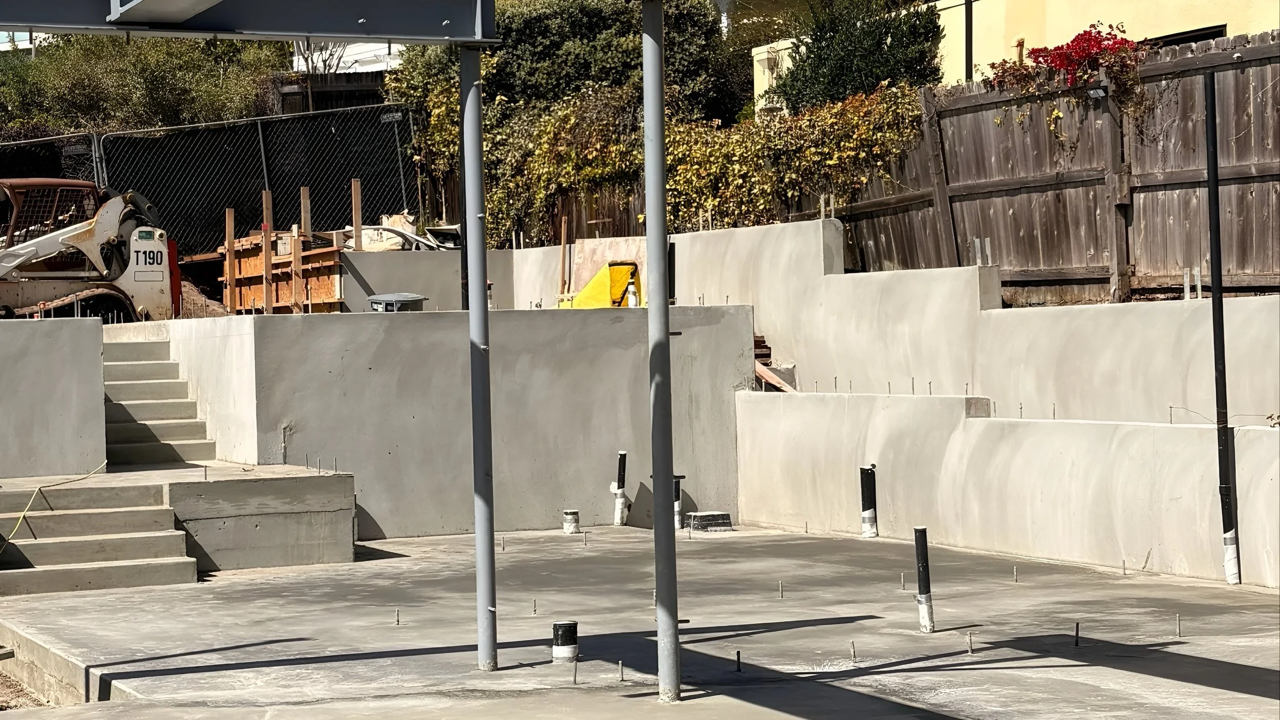 Construction site with concrete steps, newly poured concrete floor, and curved concrete retaining walls, with construction tools and materials scattered around.