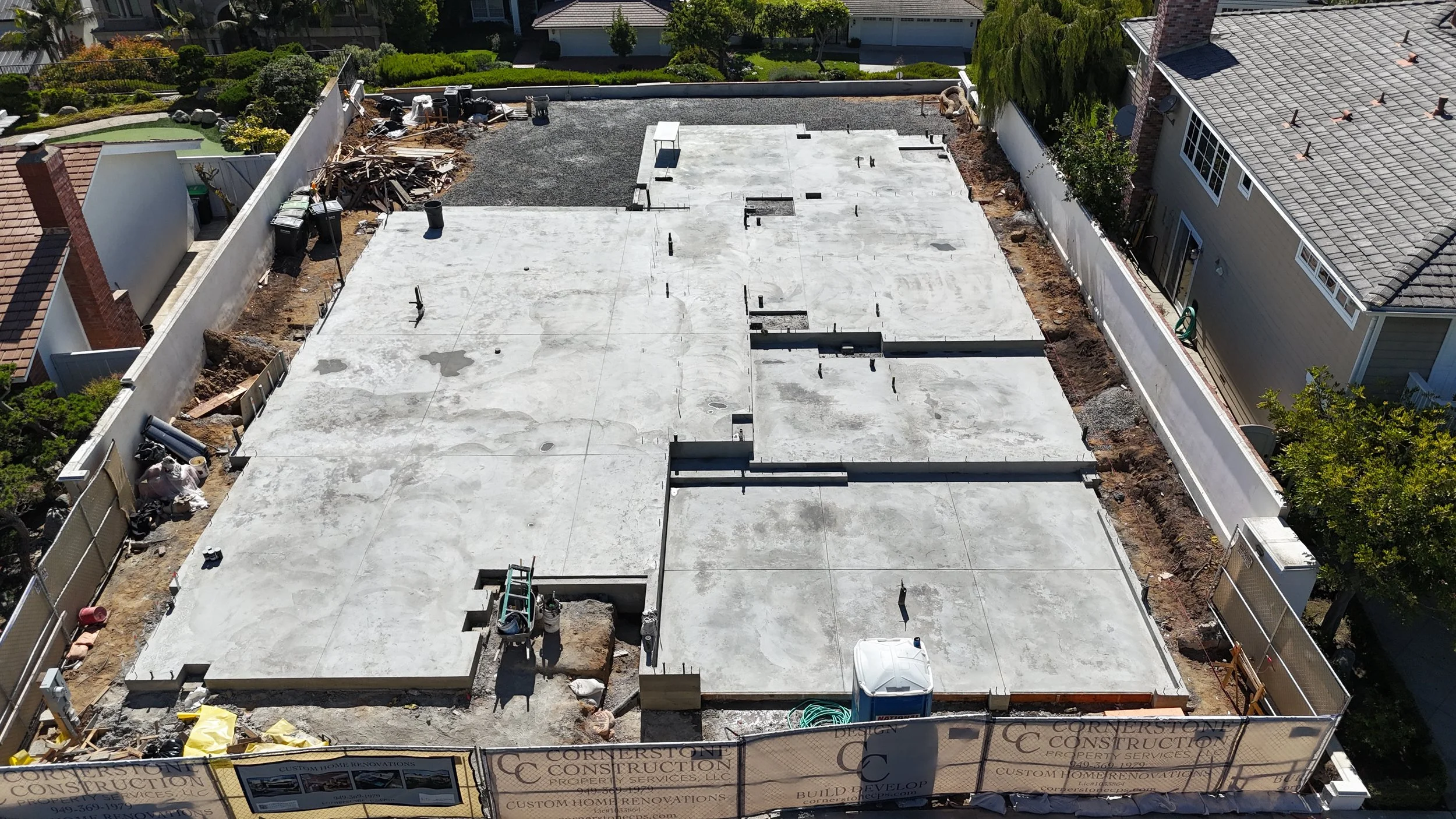 Construction site with concrete slabs in a backyard, surrounded by a white fence, with construction equipment and supplies visible.