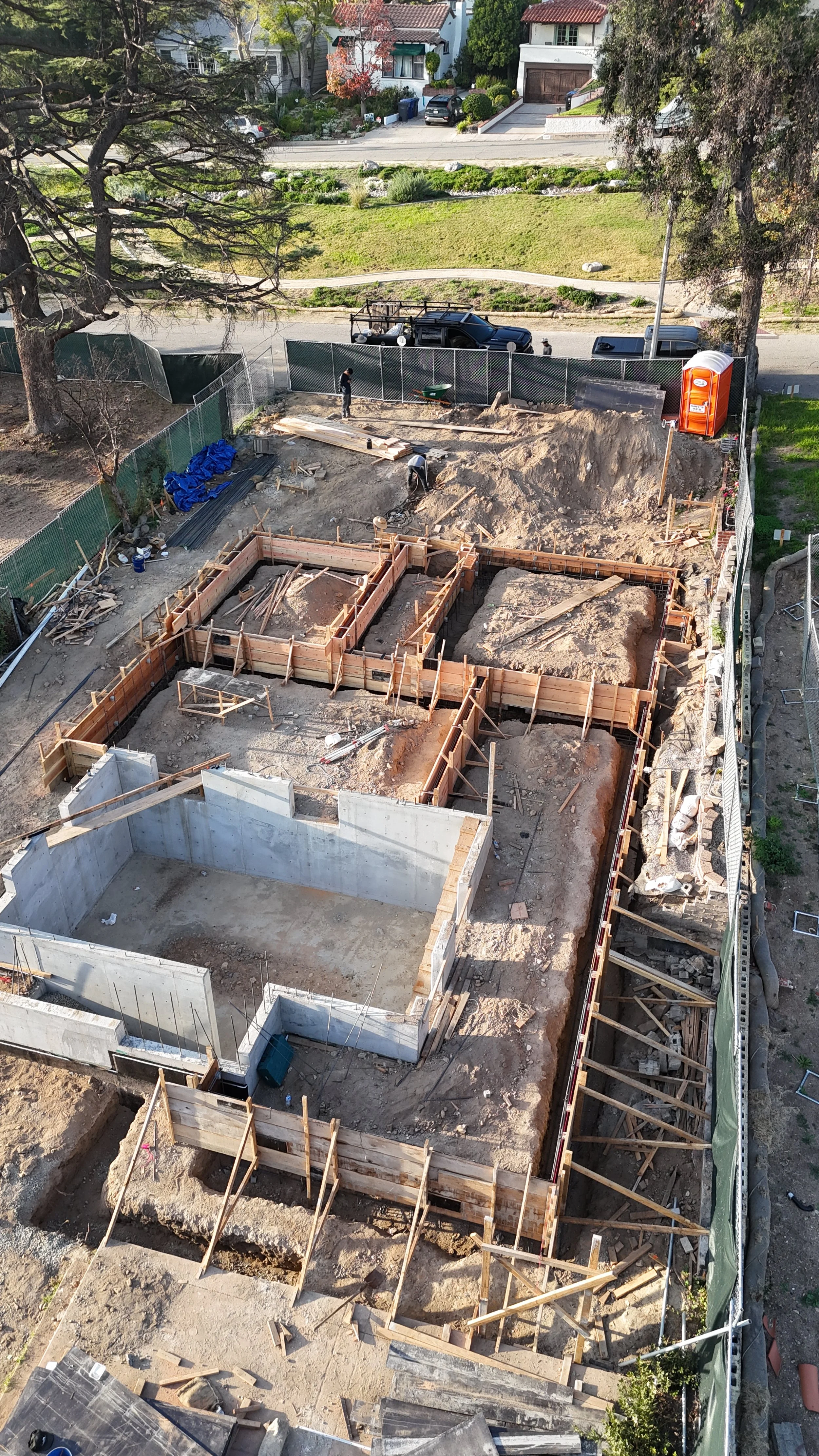 Construction site with foundation work in progress, showing concrete walls and wooden frames for future structures, surrounded by fencing and construction equipment.