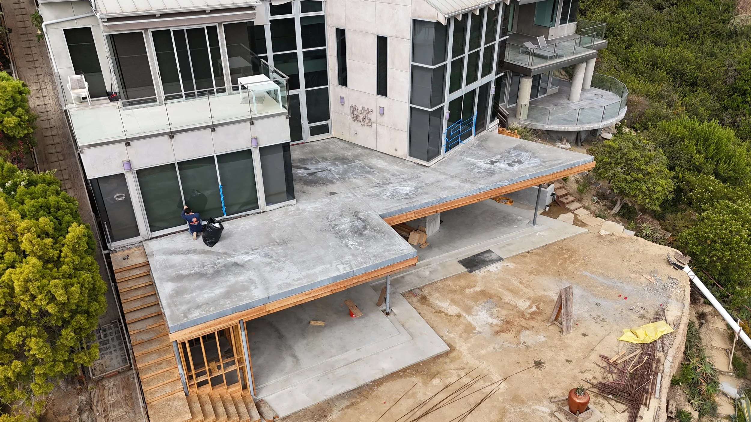Construction site of a multi-story building with concrete floors, large glass windows, and a small balcony on the upper level. There is a worker on the lower level near a black trash bag. Surrounding area has trees and construction materials.