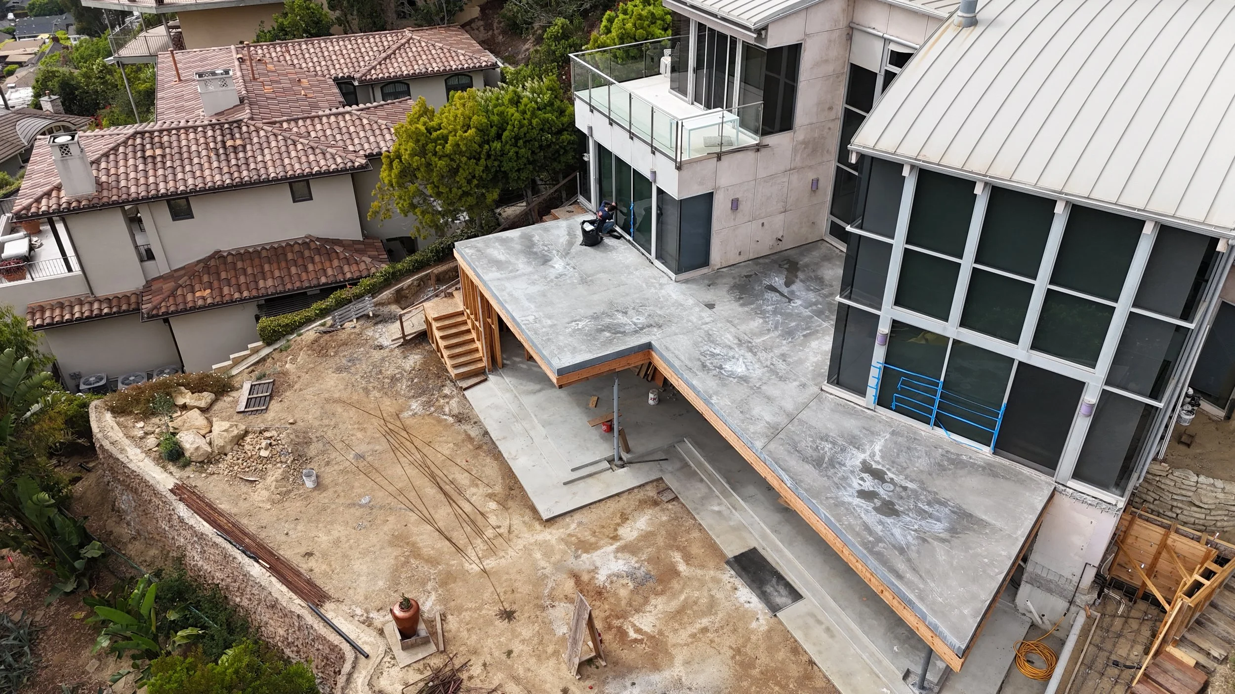 Aerial view of a residential construction site with a completed concrete patio and a partially built wooden deck, surrounded by neighboring houses with red tile roofs and trees.